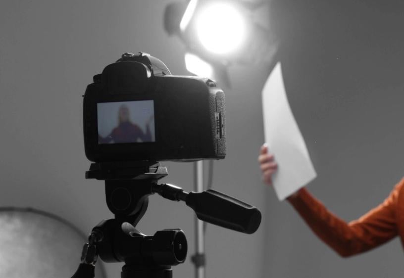 An image of a black camera on its tripod filming a woman holding a piece of paper in her hand with a light shining on her.