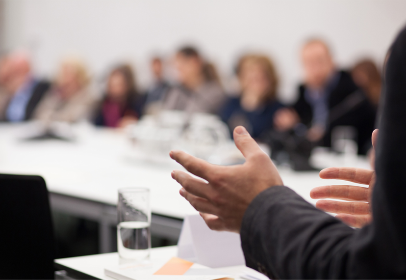 Indoor well lit meeting room with a speaker standing and attendees seated.