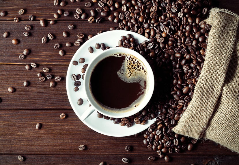 Image of a coffee cup resting on a wood table strewn with coffee bean. The cup is filled with coffee surrounded by coffee ben