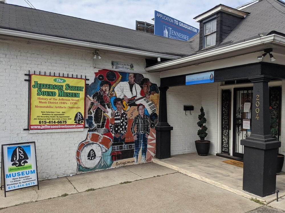 White and black building with a painted colorful mural depicting musicians playing instrumnets such as a guitar, saxophone, and drums. A sign on the wall provides information about the museum, including its hours and contact details. 