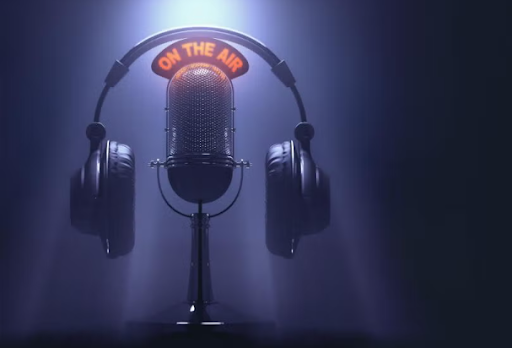 A black radio microphone stands with "on the air" in red wording above it. On top of the "on the air" sign is a pair of black headphones. The microphone, "on the air" sign and headphones appear in a spotlight in front of a black background. 