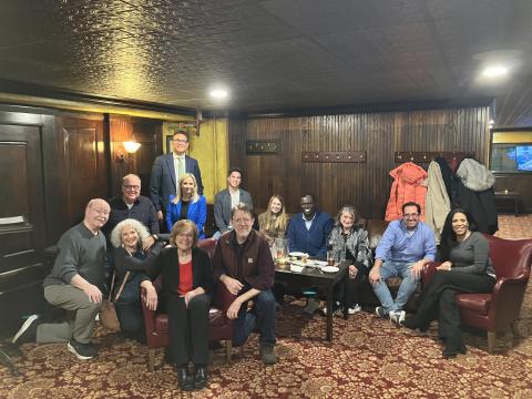 A group of smiling people, some seated on couches and chairs, some standing, smile as they pose for a photograph in a room with dark wood paneling. 