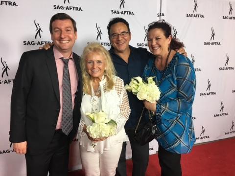 Two men and two women in business attire pose for a photo in front of a white banner with SAG-AFTRA logo.  All are smiling and happy, the two women are holding bouquets of white flowers