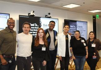 A diverse group of seven people pose for a photo in a classroom. There are three large screens behind them. 