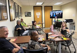 A group of diverse actors sit in a boardroom with a large screen filled with Zoom attendees at the front of the room; they smile at the camera. The carpet is standard office patterned gray, with off white walls adorned with framed movie posters. A large copier/printer/fax machine sits in the right side of the room with a trashcan near it. There are two wooden doors at the back of the boardroom.