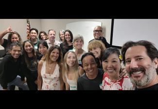 A group of diverse actors pose for a selfie in a classroom. There's an American flag in the background.