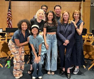 A diverse group of people pose in various clothing styles in front of directors chairs with wood paneling behind them. The carpet is blue and an American flag is on the left side. 