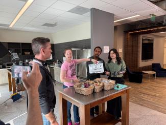 Four people are positioned around a table in a kitchen. One person is holding a clapboard in front of George. The table has baskets of food and a bright blue cutting board. On the left side, someone is recording the scene with a smartphone. 