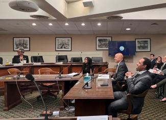 Several individuals are seated in a government chamber. George is visible in the foreground in the lower right corner of the photo. He wears a gray suit and is captured mid-laughter. 