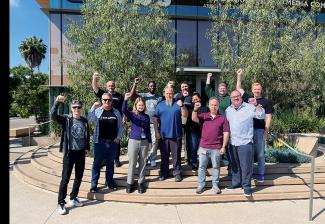 A diverse group of 12 individuals stand outside a building. Each raises a fist in solidarity.