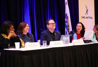 A woman with a black bob and a black and gold outfit, a woman with red shoulder length hair and a blue dress, a man with short brown hair and glasses and in a black dress shirt and navy blazer, and a woman with black hair and a red dress sit behind a black curtained table with a white top and name placards on top. 