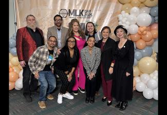 A group of individuals pose in front of a peach backdrop with peach, pale yellow, pale blue and white balloons on both sides.