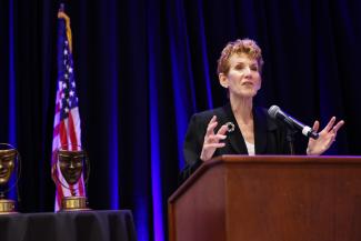 A woman with a short red hair and black blazer with a white top speaks behind a wooden podium with a mic. A dark blue curtain is draped behind her with an American flag in the left of the background. Two theater mask awards sit on top of a black tablecloth in the bottom left of the photo.