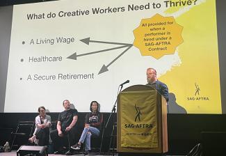 A man with a beard and a ponytail stands at a podium with a yellow SAG-AFTRA banner on the front of the podium. A projection screen hangs behind the podium showing a presentation. The slide on the screen asks “What do Creative Workers Need to Thrive?” Next to the podium, three panelists sit in chairs. One panelist is a woman with brown hair who wears a long white shawl. The man in the middle is wearing a black T-shirt and black pants. The third woman wears jeans and a black SAG-AFTRA Pride shirt.