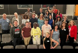 A diverse group of people pose for a photo among the seats in a theater. Most of the people wear casual clothing and a nametag.