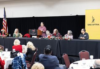 A person stands at a podium speaking, while six others sit on the dais on either side. People sitting at round tables listen to the speaker.