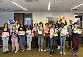 A group of people stand in a row holding pieces of art they have made out of canvas and multicolored pieces of paper.