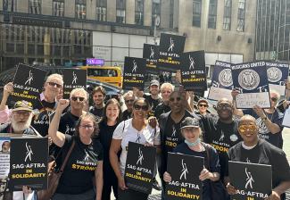 A group of people are holding black signs with the white SAG-AFTRA logo, reading “SAG-AFTRA in Solidarity” in white and yellow text. There are people to the right of the image with navy and white signs that read “Musicians United: Better Healthcare.”    