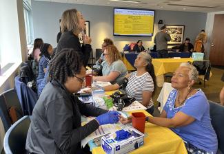  A group of people sit on two sides of a table covered with a yellow tablecloth. The people on the left side perform finger sticks and blood pressure readings on the people on the right side.