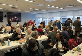 A large, diverse group of people seated in rows at tables inside a room. There are plates of food on the table. A yule log video appears on the screens in the background.