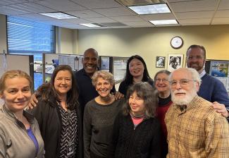 A diverse group of people smile in front of cubicles and a yellow wall with a clock and framed pictures. 