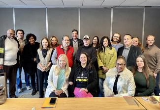 A diverse group of people pose for a photo behind a conference table and in front of a gray wall at the conclusion of a meeting.