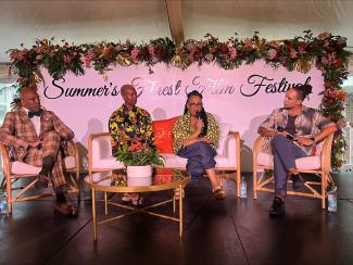 From left, Knight, Smith, Price and Maslen, are seated on stage. Price speaks with her microphone held closely to her mouth as the others look on intently. In the foreground is a table with flowers; behind them is a lavender wall with “Summer’s Finest Film Festival” in cursive writing and framed with flowers. 