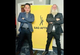 Two people in suits stand with their arms folded on either side of a yellow SAG-AFTRA banner.