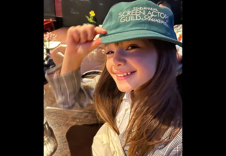 Quinonez, a young girl with long brown hair, smiles at the camera. Her right hand pinches the brim of a green sports hat with the copy “22nd Annual Screen Actors Guild Awards” in gold thread.