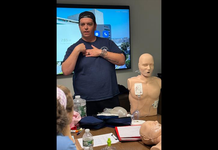 A man wearing a backward baseball cap, a blue T-shirt and khaki pants stands at the front of a classroom. He demonstrates how to use an automated external defibrillator. There is an adult CPR mannequin placed on a table.