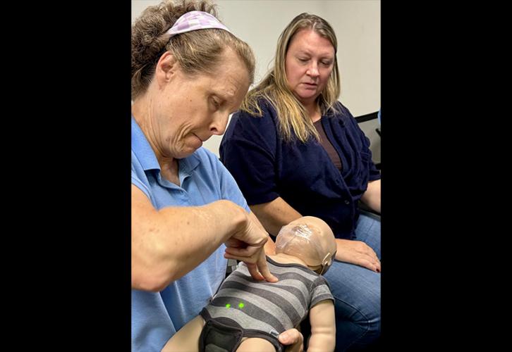 A seated blonde woman in a light blue short-sleeve collared shirt with a cloth headband presses two fingers against an infant mannequin that lays on her lap. A second woman with long blonde hair dressed in blue observes.