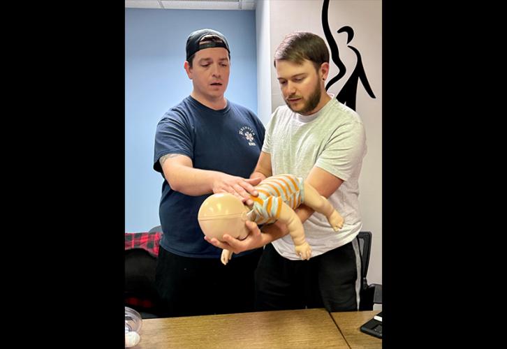 A man wearing a backward baseball cap, a blue T-shirt and khaki pants aids a second man holding an infant mannequin. The second man wears a white T-shirt and black pants.