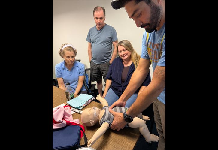 A man in a blue T-shirt and khaki pants holds an infant CPR mannequin he has placed on a table around the chest. Three additional people, two women and one man, observe the practice. One woman, seated, wears a light blue short-sleeved collared shirt with a cloth headband. A man, standing, wears a gray T-shirt and dark gray shorts. The second woman has long blonde hair and is dressed in blue.