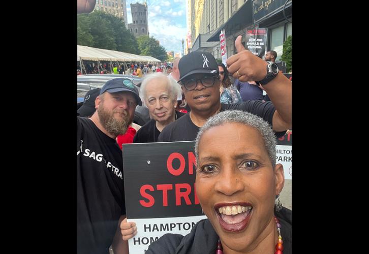 A woman with short gray hair is in front and the center of a selfie with three men behind her on a busy street. All four people are wearing black. The first man has a beard and is wearing a baseball cap, the second man has gray hair and the third man, wearing glasses and a black baseball cap, is giving the thumbs-up sign with his left hand.