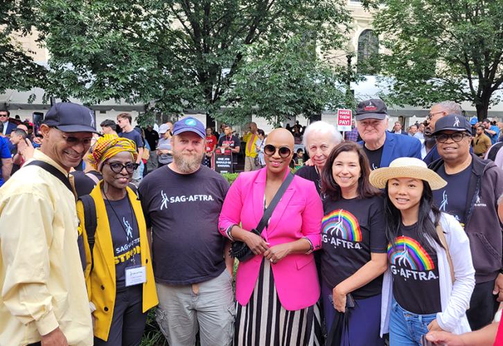 A diverse group of people stand on either side of Rep. Ayanna Pressley, center, in an outdoor area. Pressley wears a black and white striped dress with a pink blazer and a black crossbody purse. Three people stand on one side of her and five people stand on the other side, all wearing a variety of SAG-AFTRA T-shirts.