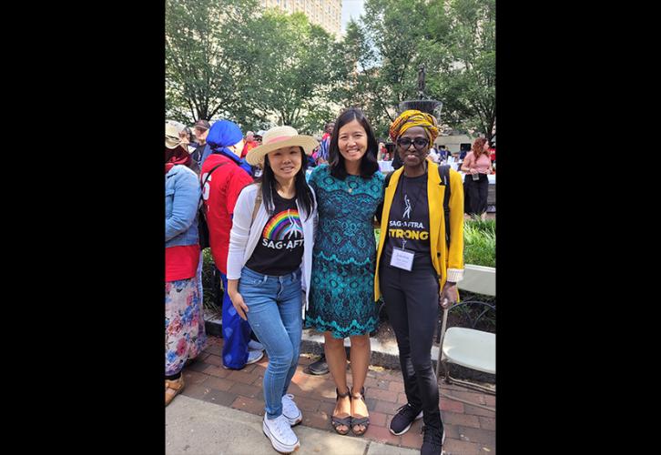 Wu, wearing a teal and black patterned dress, stands in the middle of a group of three women with each of her arms on the shoulder of a woman on either side of her. The woman on her left wears a colorful hair wrap, a yellow suit jacket, black pants and a black “SAG-AFTRA Strong” T-shirt. The other woman wears a straw hat with a pink ribbon, a black SAG-AFTRA Pride T-shirt with a rainbow and an open white button-down sweater with jeans.