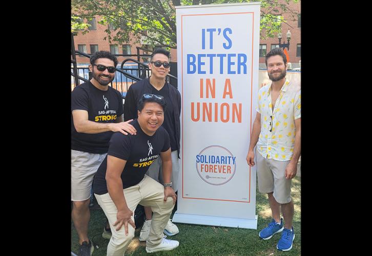 Three men, all in black shirts and tan pants/shorts, with two of them wearing sunglasses, stand on one side of a tall vertical white banner that reads in blue and orange letters “It’s Better in a Union.” A man with a beard stands on the other side of the banner wearing tan shorts, and a white and yellow top. Sunglasses hang from his shirt.