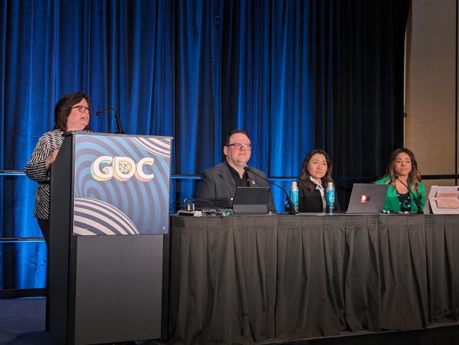 Oratz, left, stands behind a podium with GDC 2024 artwork. Crabtree-Ireland, Henderson (African American) and Wang (Asian American) sit at a table with a long black tablecloth on stage.