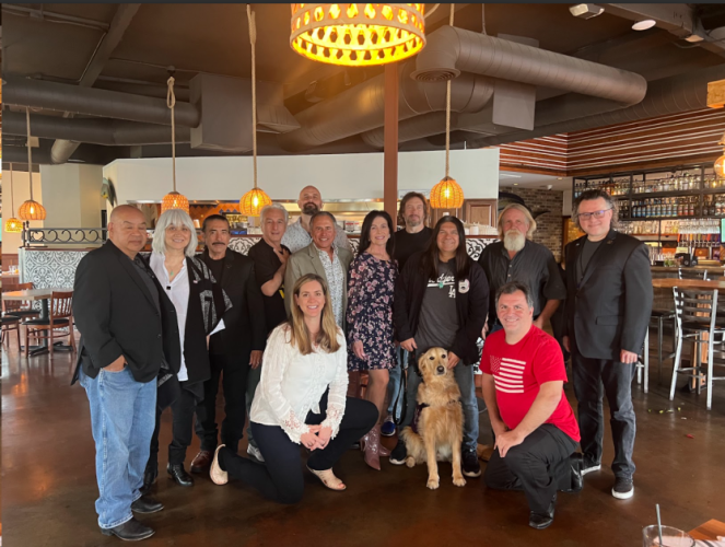 A group photo of 13 individuals in a restaurant area. A woman and a man in a red shirt are kneeling. There is also a dog.