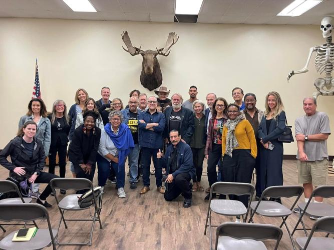 A group photo of a couple dozen people taken in a brightly lit room. In the foreground are rows of metal chairs; to the right is a large skeleton; and there is a moose head above the group.