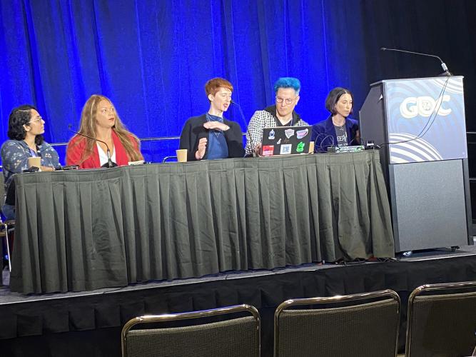 Kunapaneni, Schermerhorn, Elmaleh, Karliak and Rousseau are seen behind a table and podium on stage. Schermerhorn wears a distinctive red blazer; Karliak’s hair is blue. Elmaleh speaks to Karliak.