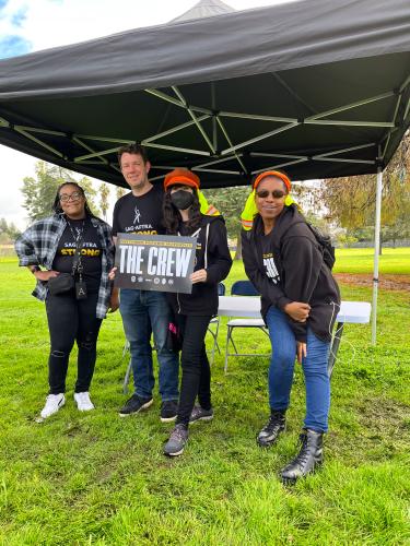 Four individuals, three women and one men, stand in the grass under a tent with a black tarp. They are all dressed casually in SAG-AFTRA Strike t-shirts and jeans. 