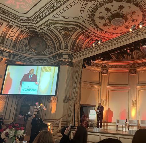 Knight (African American) stands behind a podium on stage. On a projector, left, he appears in a front profile. Also seen is the architecture of the room, notably the gold fixtures on the wall. 