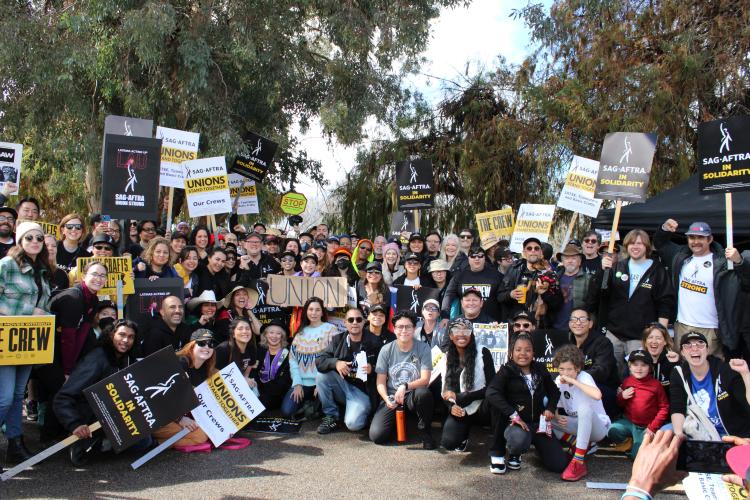 A group photo of several dozen individuals arranged in multiple rows. Those in front kneel on the ground. Visible throughout are picket signs, including SAG-AFTRA Strike signs.