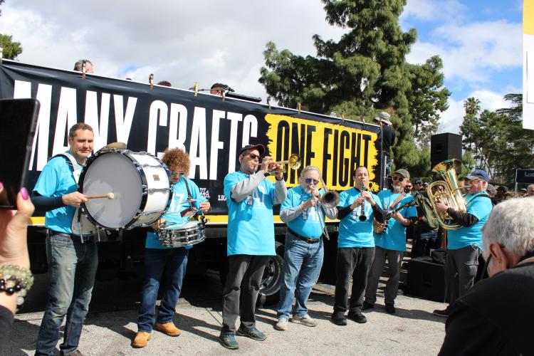 Seven individuals play instruments including horn and percussion instruments. They all wear sky blue t-shirts bearing the AFM Local’s logo. 