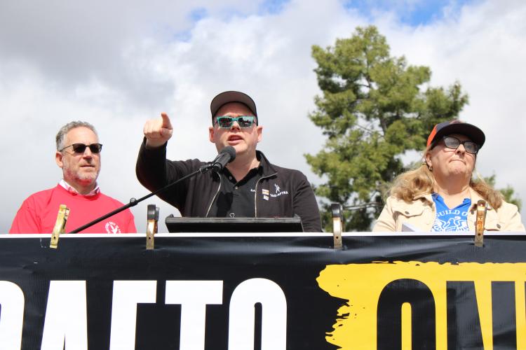 Crabtree-Ireland stands onstage and speaks into a podium with a mic. The rally banner hangs across the front and is obscured. On his either side is a man in a red shirt and a woman in a blue shirt with a khaki jacket. All wear sunglasses.