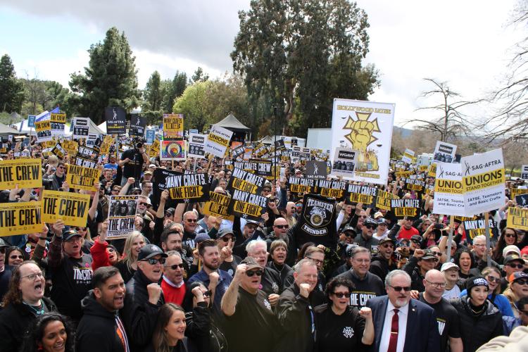 A crowd shot of dozens of individuals facing the stage. Those in the foreground, leaders from the various unions, raise their fists in solidarity. Picket signs from SAG-AFTRA, IATSE, The Crew and other unions are held above head.