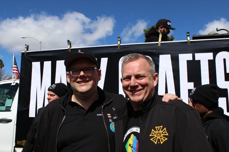 Crabtree-Ireland, left, and Miller stand closely for a photo. Both wear all-black outfits with Miller wearing an IATSE jacket and a t-shirt bearing a solidarity fist in green, yellow, blue and purple. Shadows are casted across both men’s faces. 