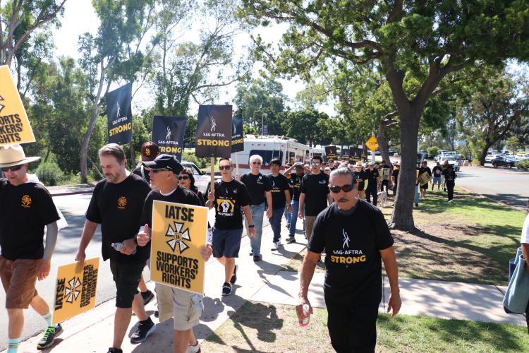 A shot of demonstrators along a sidewalk. To the left, protesters wear black T-shirts bearing SAG-AFTRA and IATSE logos, and signs from both unions can be seen throughout. Also visible are trees and foliage.