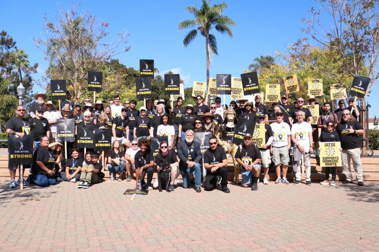 A large group photo with SAG-AFTRA and IATSE members in the park. SAG-AFTRA and IATSE picket signs are seen throughout. A brick pathway in the foreground; trees are seen in the background.